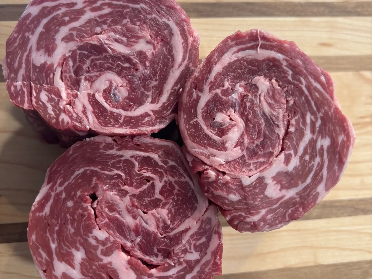 Three raw ribeye cap steaks rolled into pinwheels on a wooden board