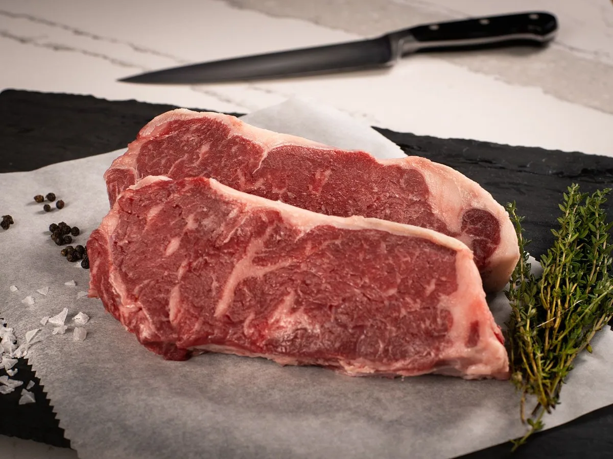 Two raw New York strip steaks on butcher paper with thyme and peppercorns