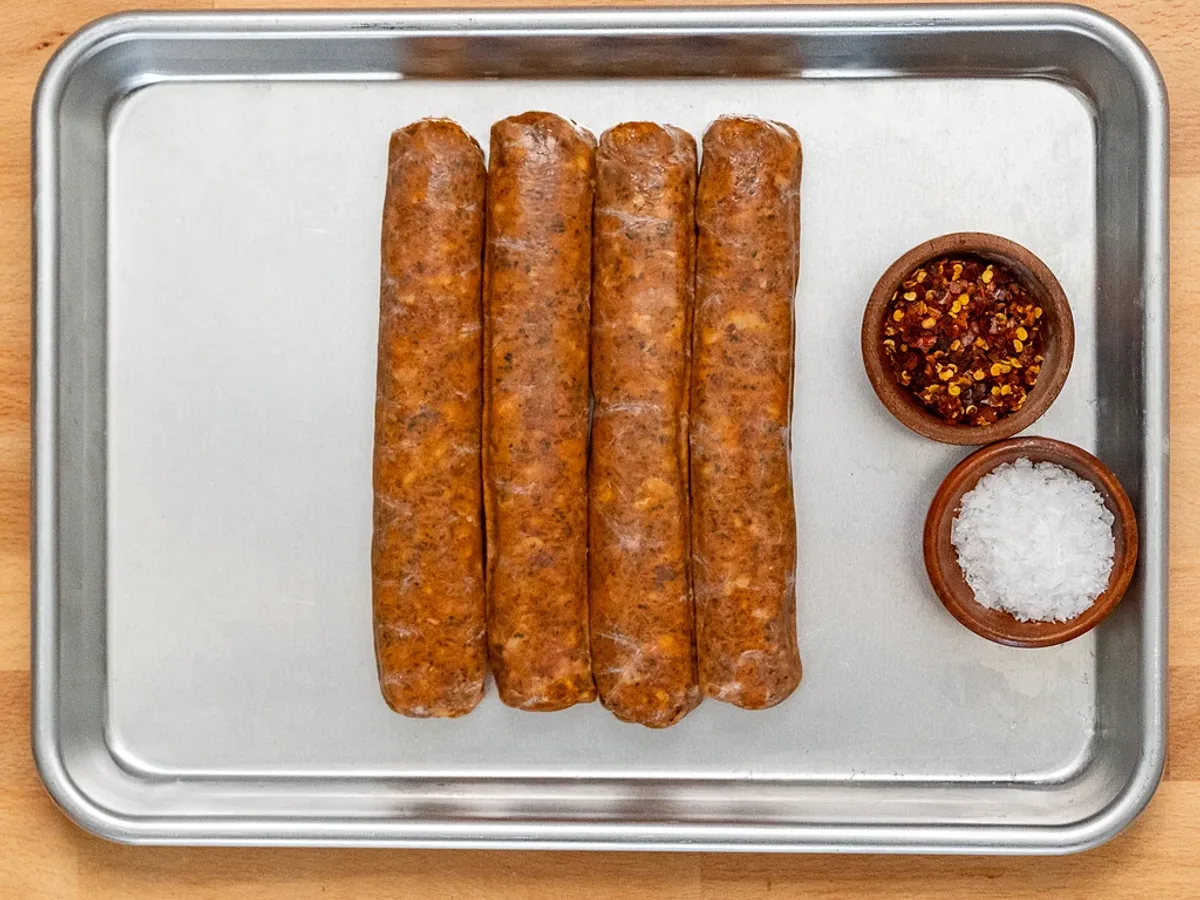 Four raw Italian sausage links on a sheet pan with chili flakes and salt